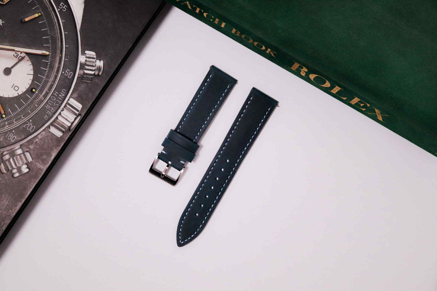 Watch strap on a white surface with a Rolex watch and book in the background