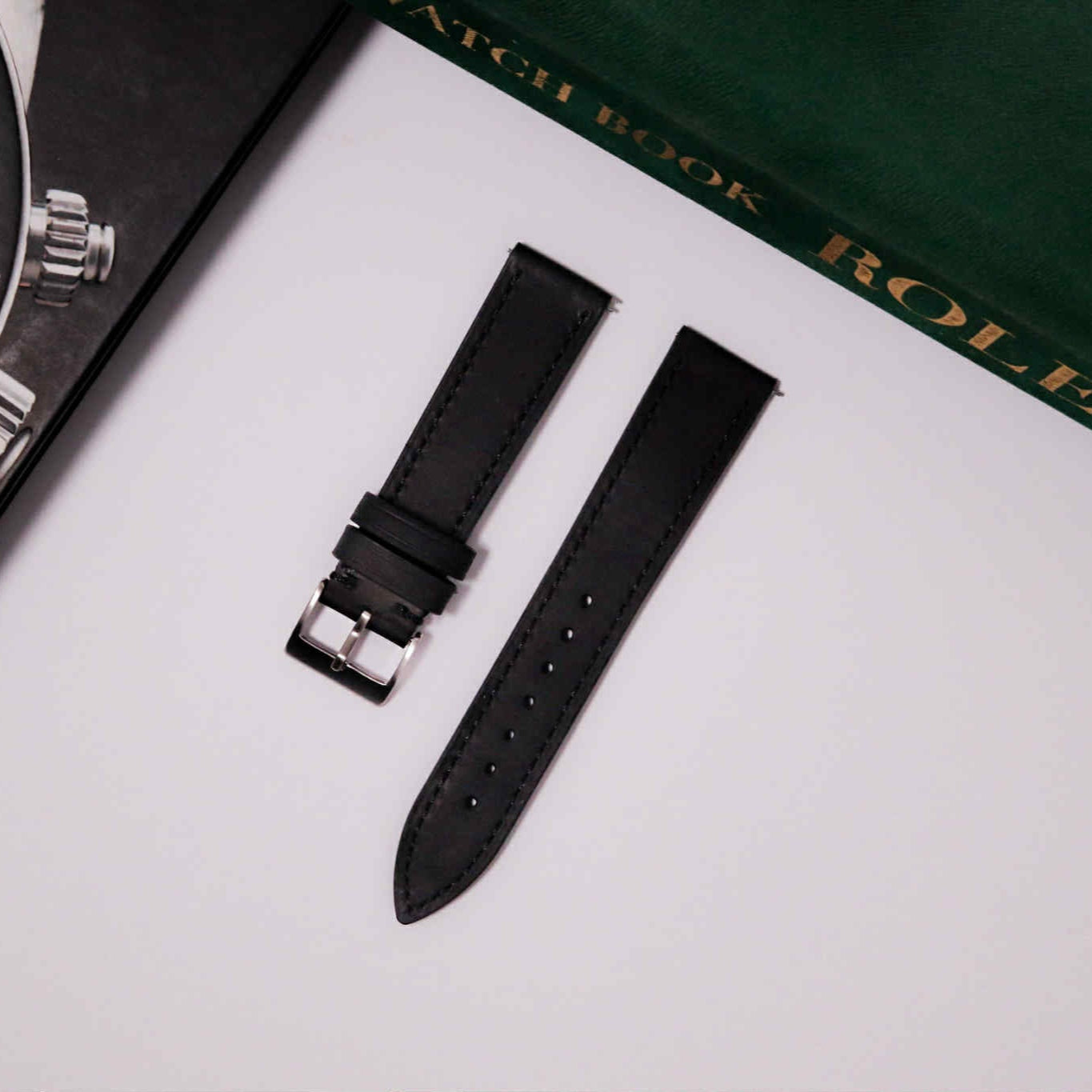 Black watch strap and watch on a white surface with a green Rolex book in the background