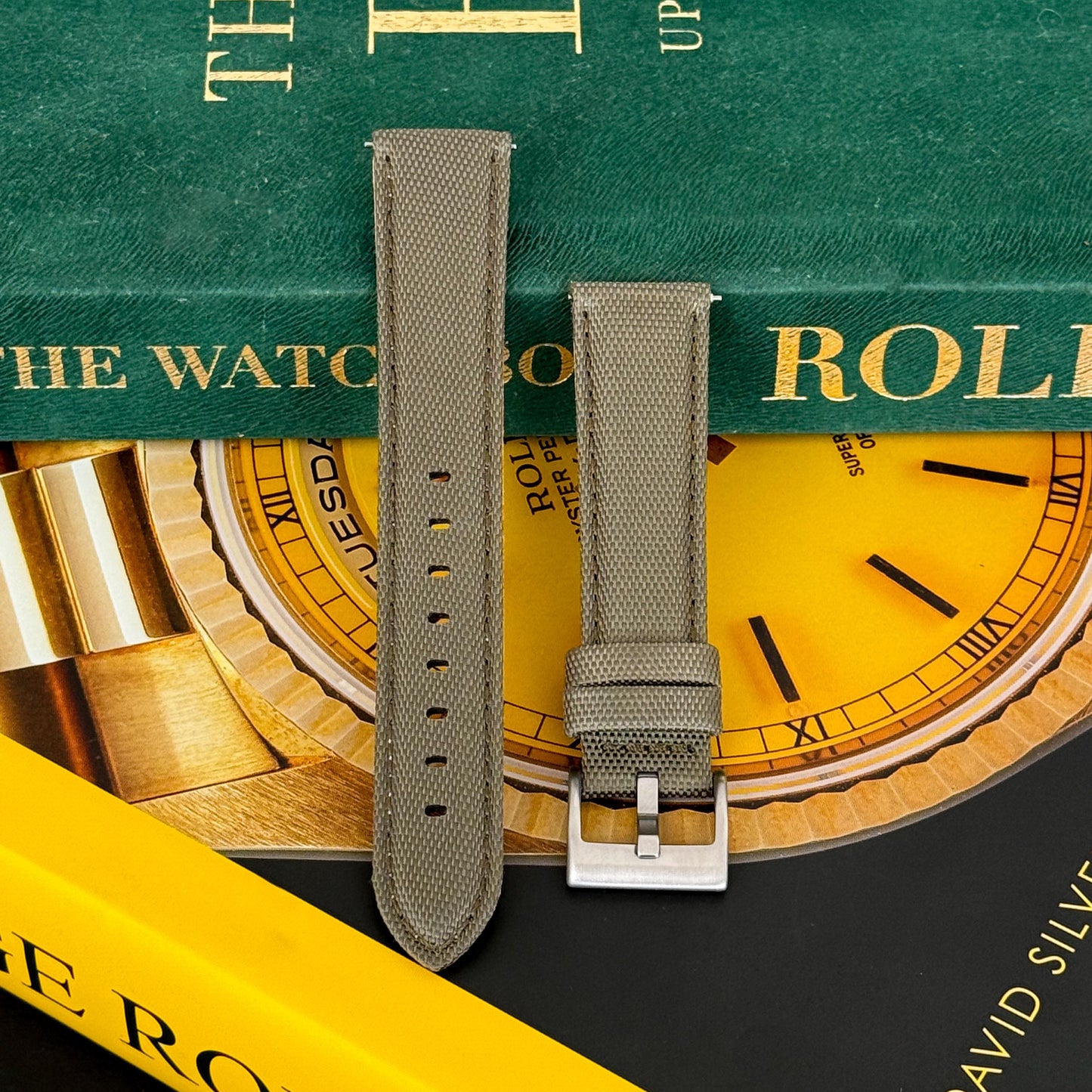 A khaki green sailcloth watch strap with a premium leather lining and a brushed stainless steel buckle, displayed on a watch-related backdrop.