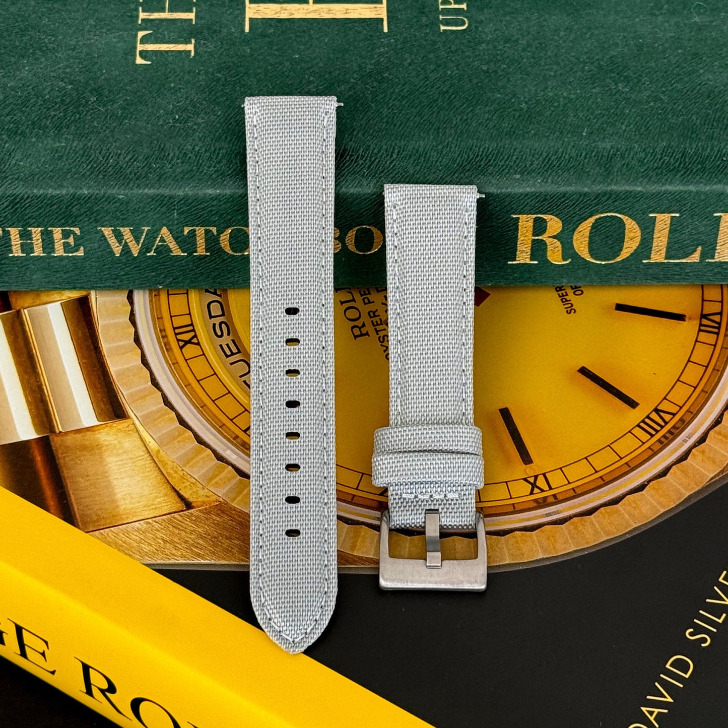 A pewter grey sailcloth watch strap with a brushed stainless steel buckle, displayed on a watch-related backdrop.