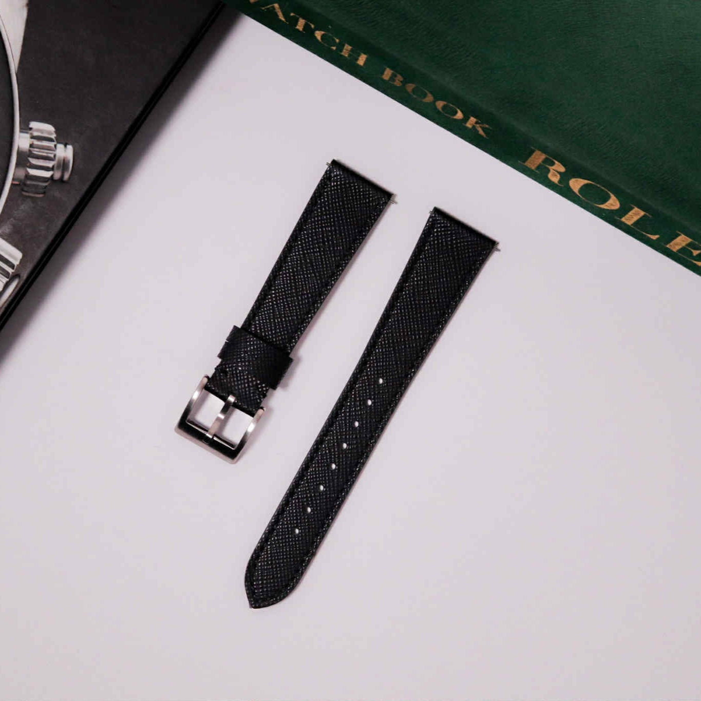 Black watch strap on a white surface with a green and gold 'Rolex' book in the background.