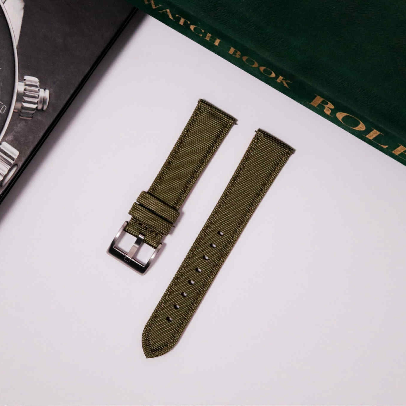 Green watch strap with a silver buckle on a white surface, next to a Rolex watch and book.