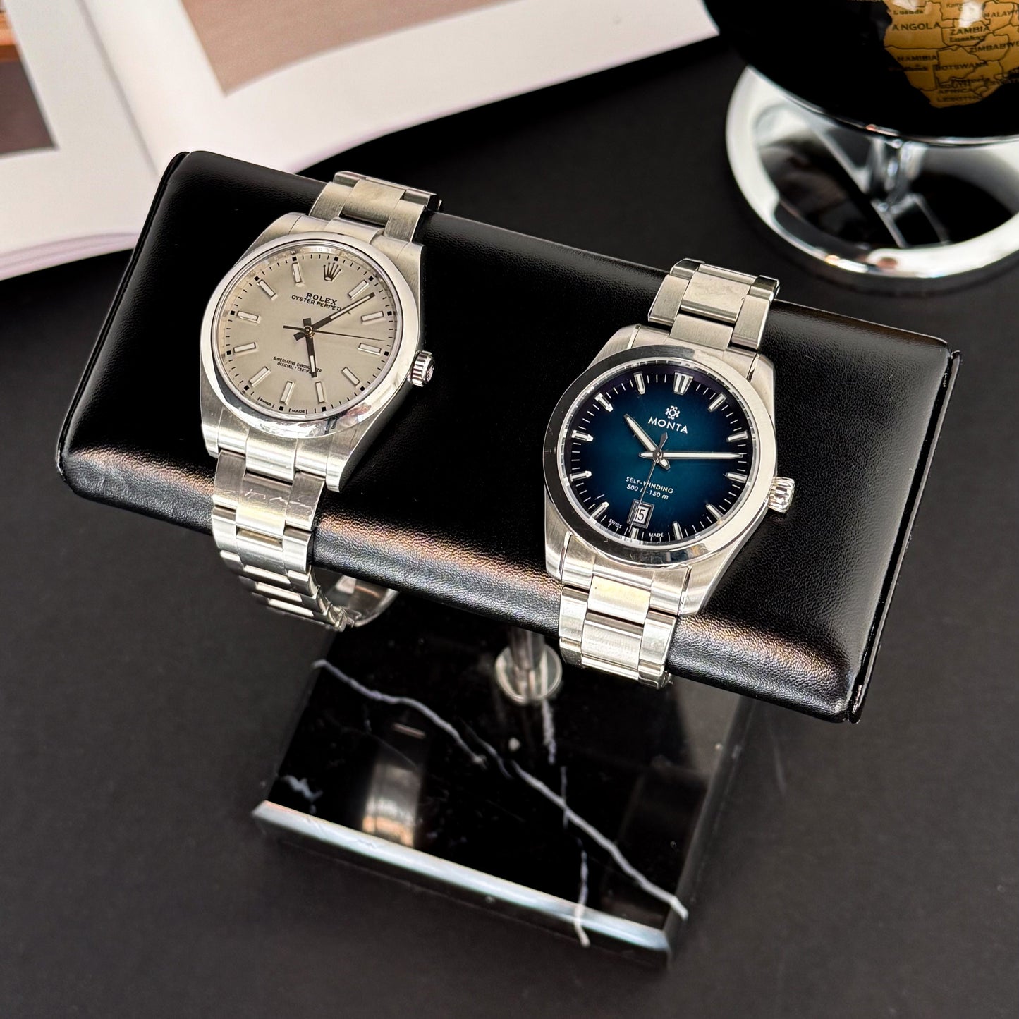 Two analog watches displayed on an m and son black double leather watch stand with a marble surface below.
