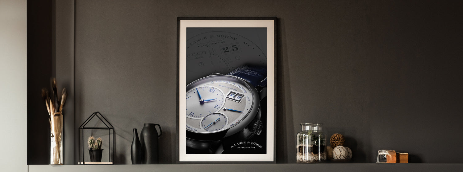 Framed artwork of an a lange watch on a dark wall with decorative items.
