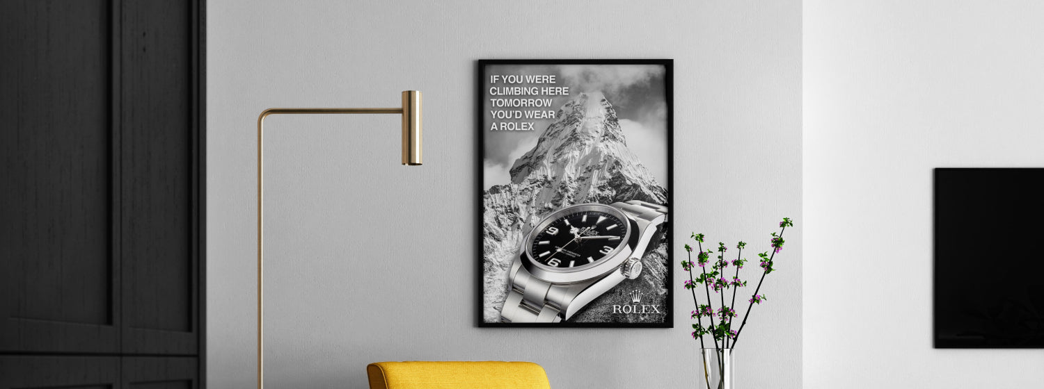 Framed Rolex advertisement on a wall with a lamp and plant in the foreground.