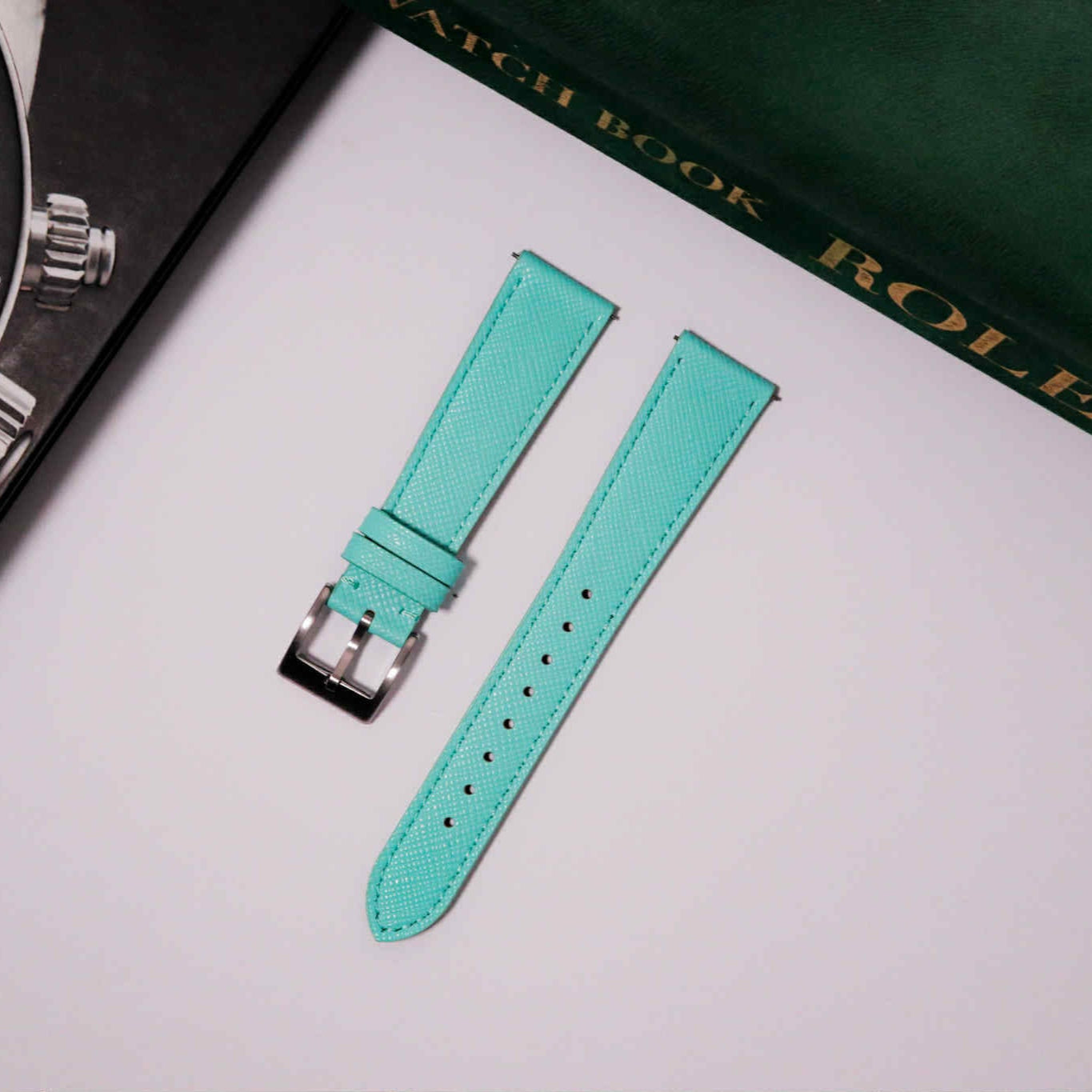 Turquoise watch strap with silver buckle on a white surface with a Rolex book in the background.