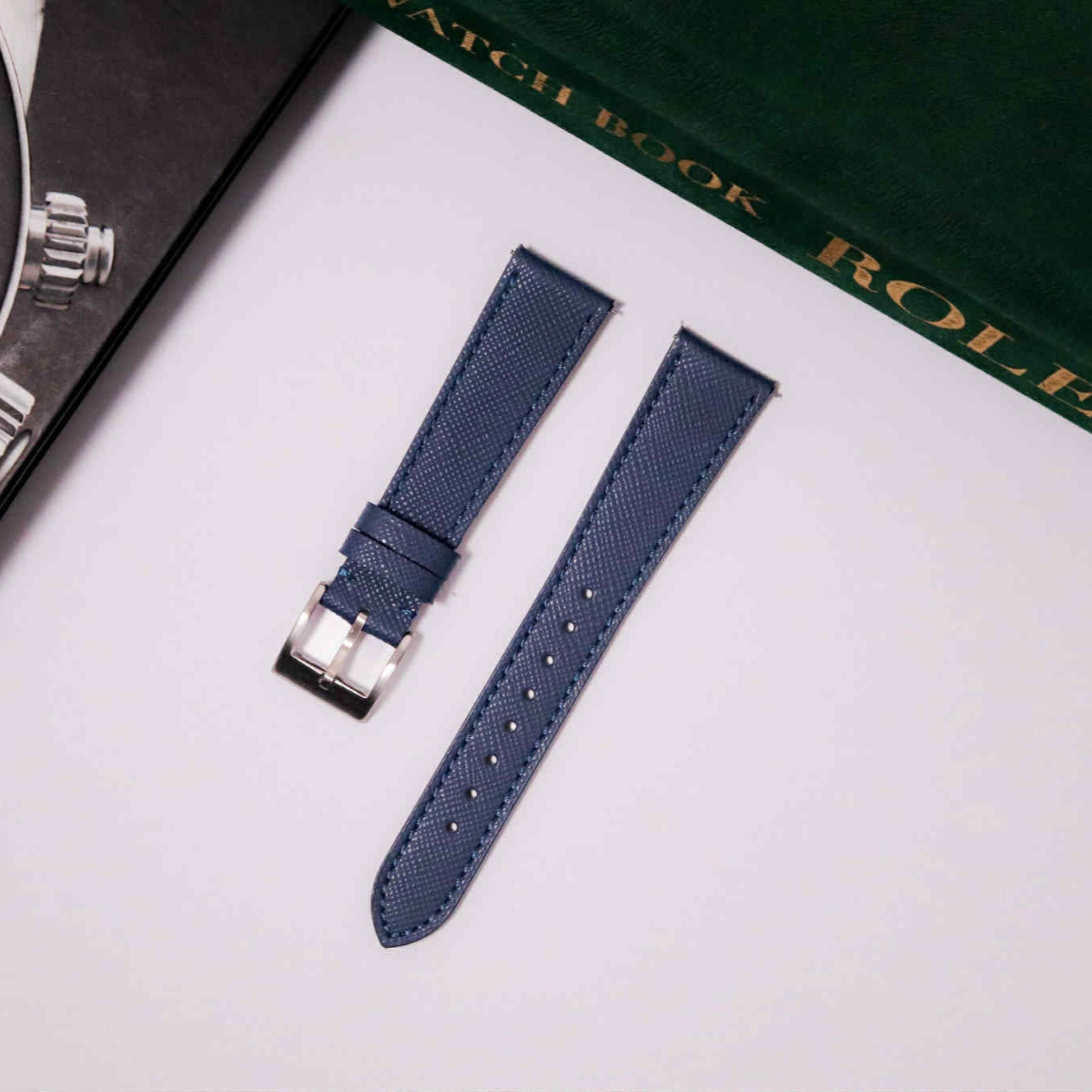 Blue watch strap with silver buckle on a white surface with a Rolex box in the background.