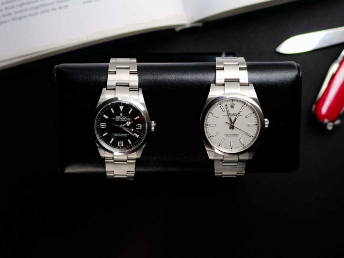 Two rolex watches on a black stand with a book and knife in the background