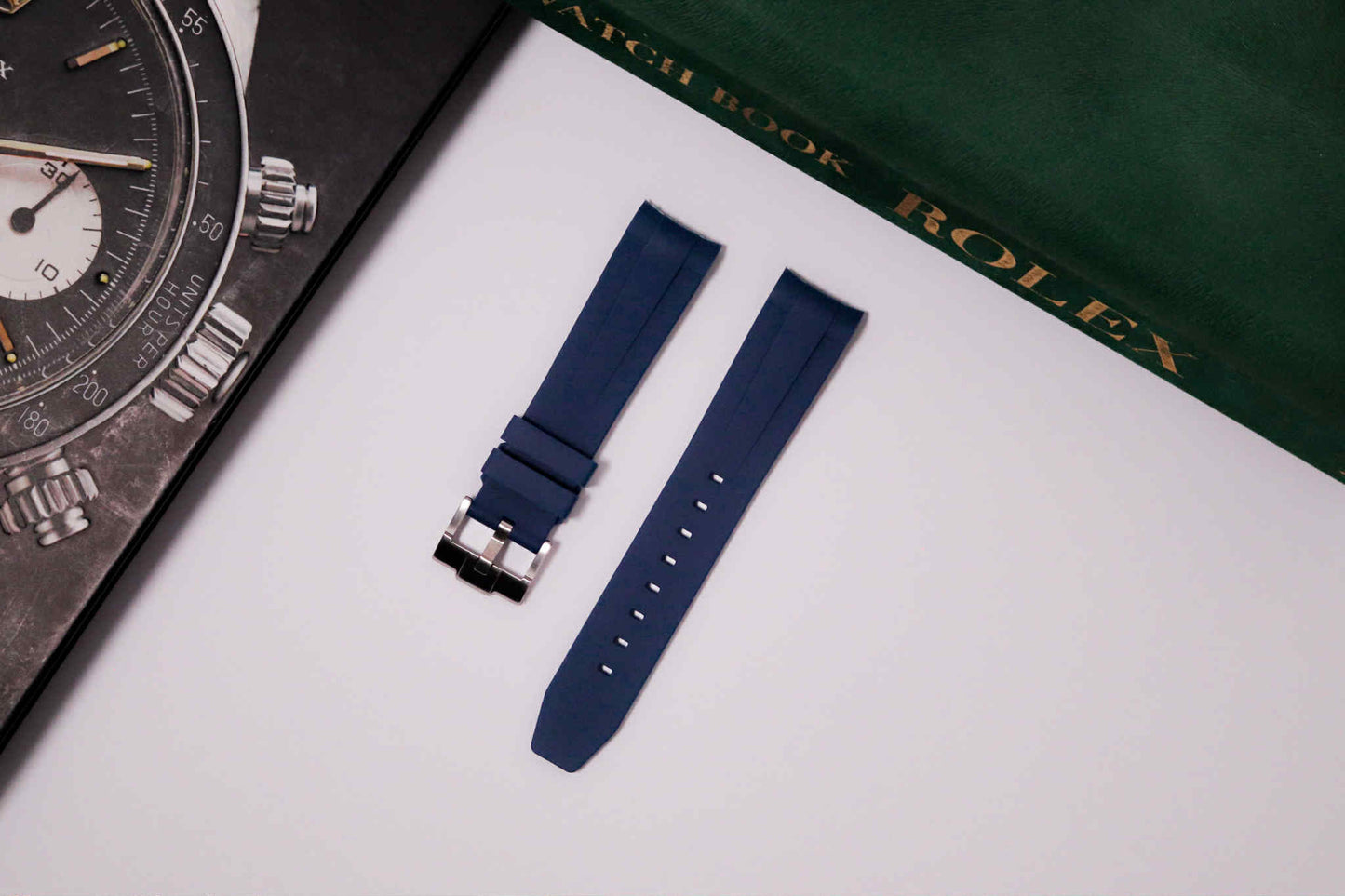 Blue watch strap with a partially visible watch and a Rolex book in the background.