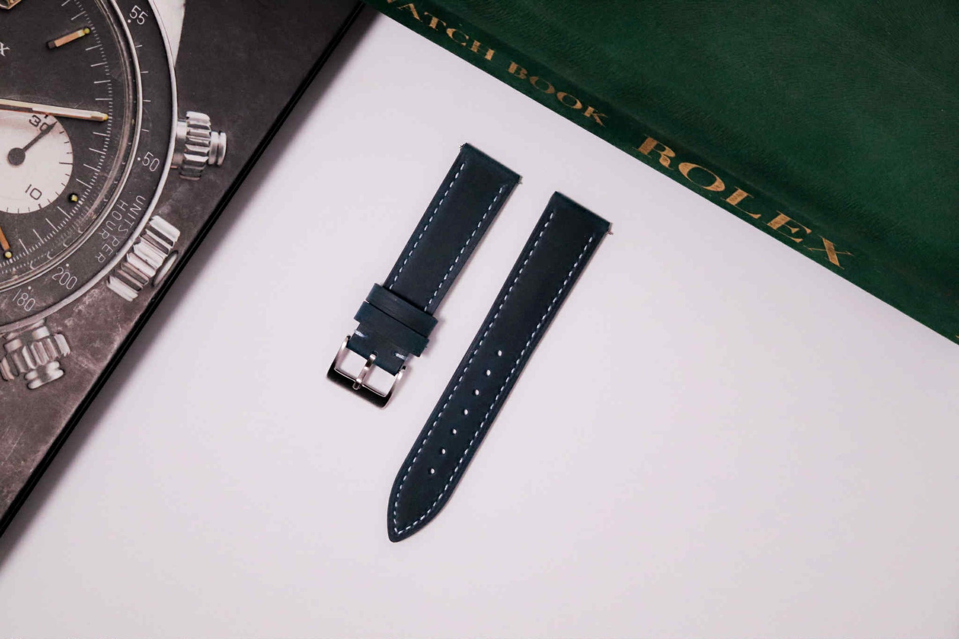 Watch strap on a white surface with a Rolex watch and book in the background