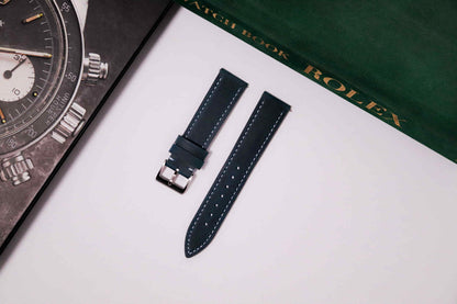 Watch strap on a white surface with a Rolex watch and book in the background