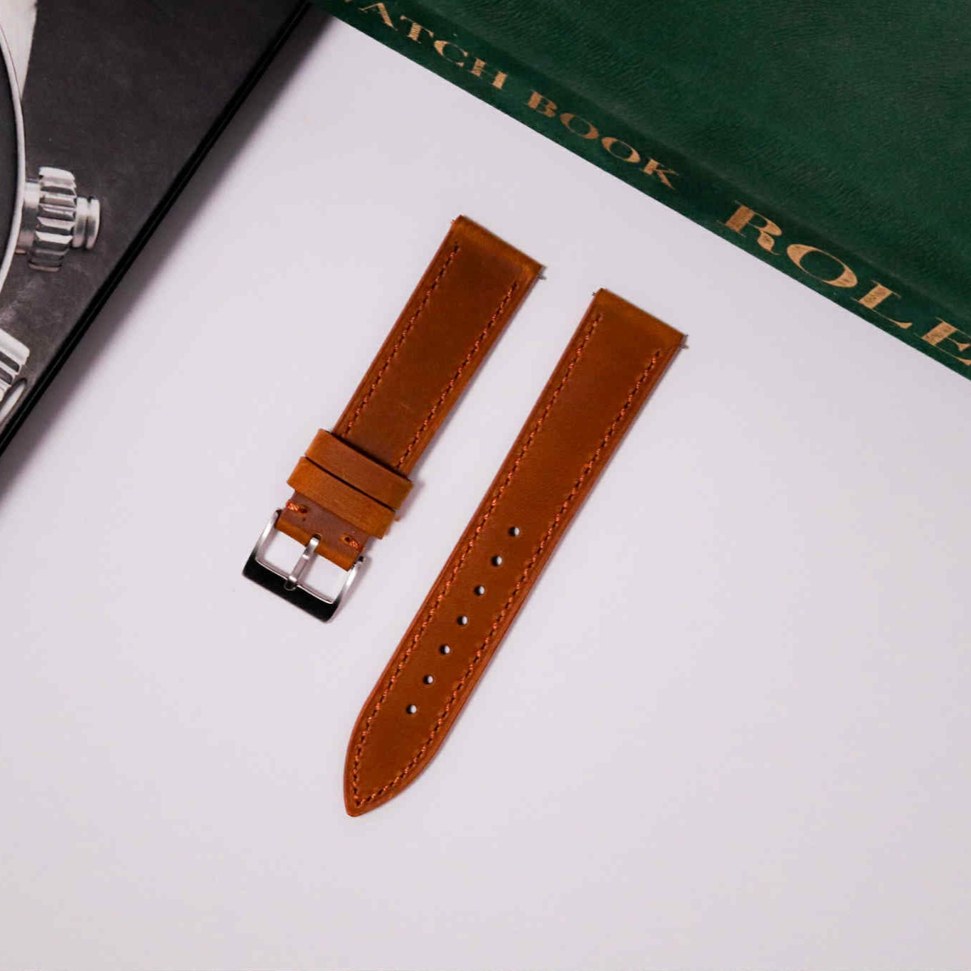 Brown leather watch strap with silver buckle on a white surface with a Rolex watch book in the background.
