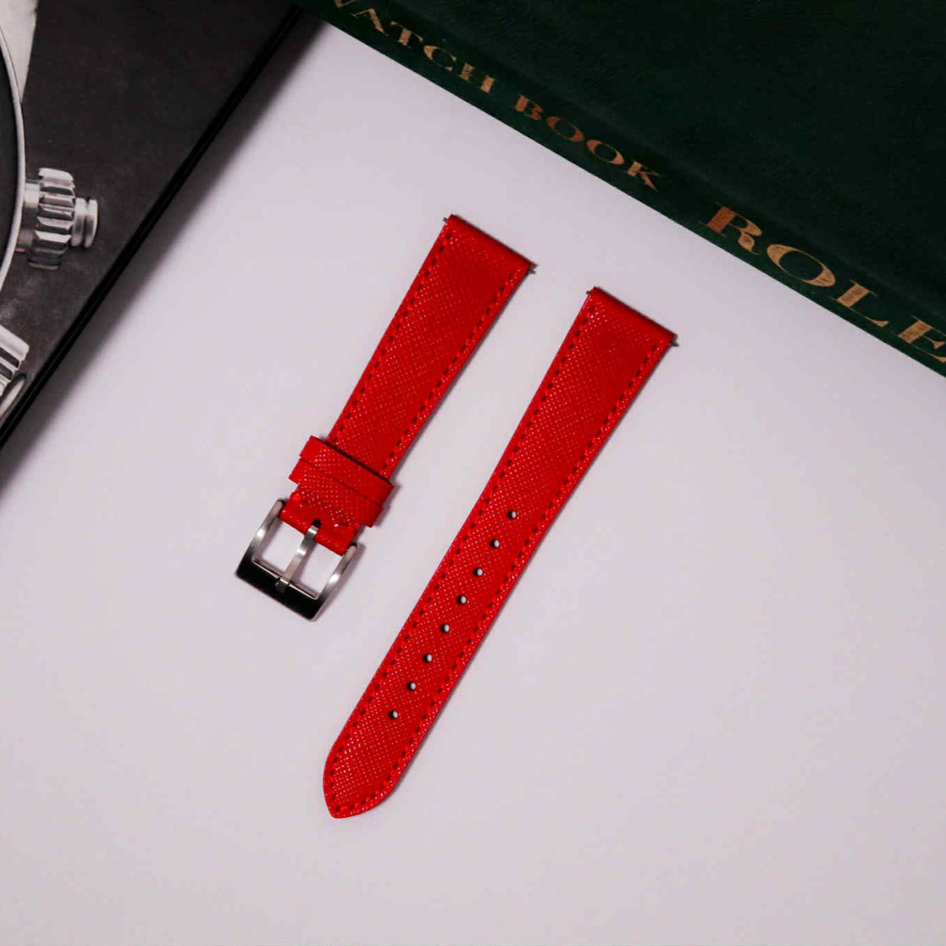 Red watch strap with silver buckle on a white surface, with a Rolex watch book in the background.