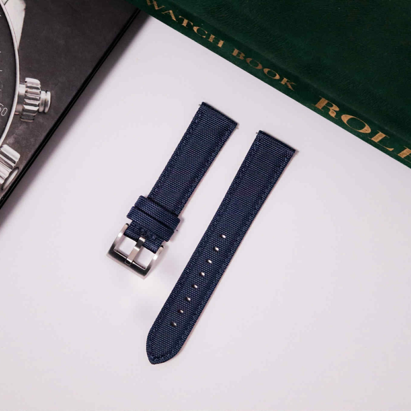 Navy watch strap with silver clasp on a white surface next to a Rolex watch book.