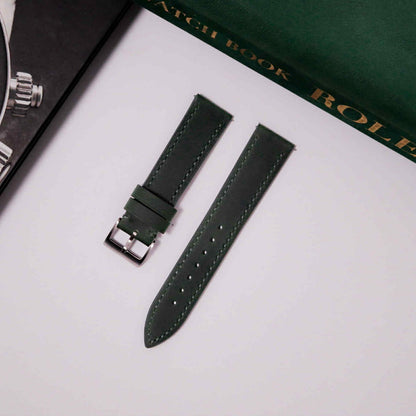 Green watch strap on a white surface with a black watch and green box in the background.