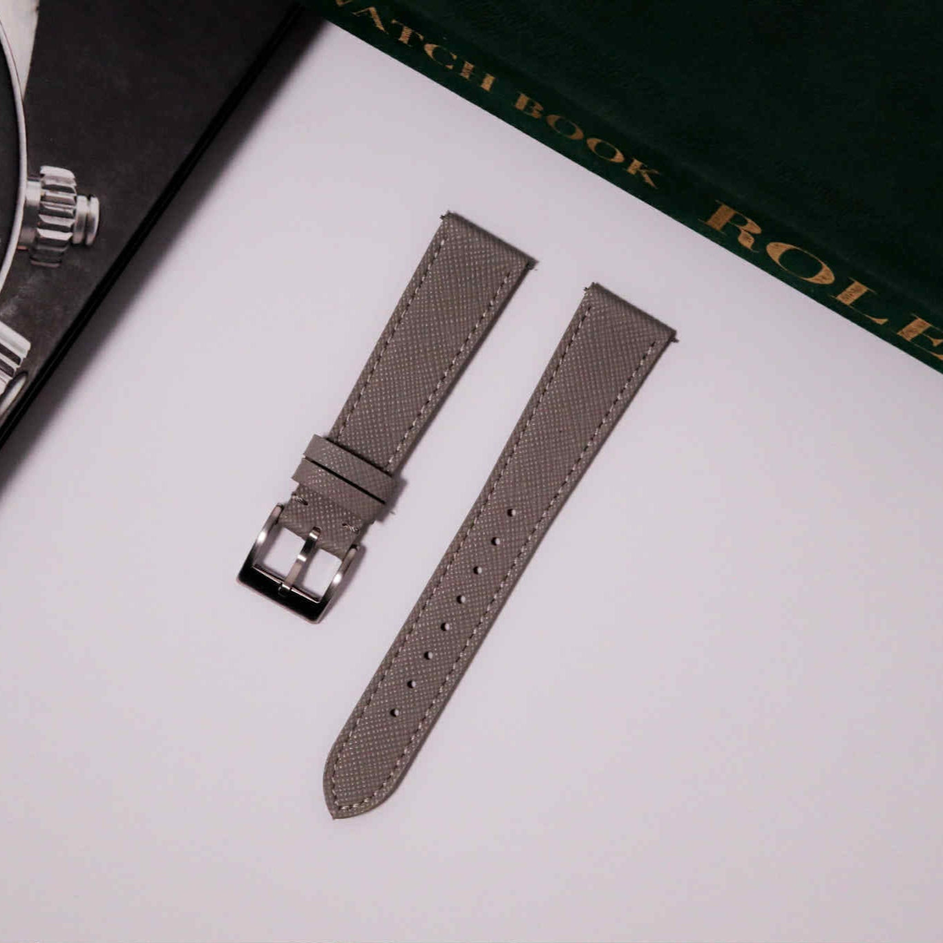 Gray watch strap with silver buckle on a white surface with a Rolex box in the background.