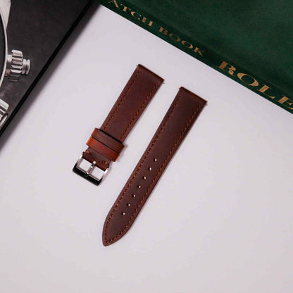 Brown leather watch strap with silver buckle on a white surface with a Rolex book in the background.