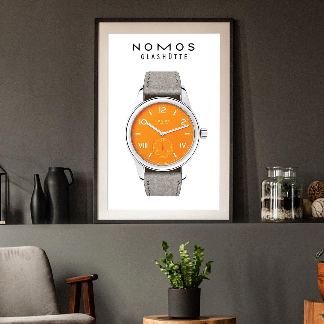 Framed poster of a Nomos Glashütте watch with an orange face and gray strap on a wall.