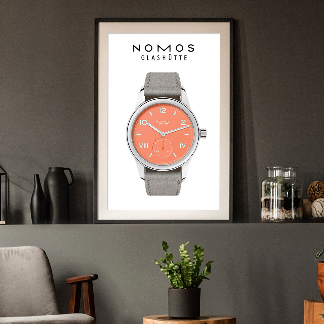 Framed poster of a Nomos Glashüte watch with a gray strap and coral face on a dark wall.